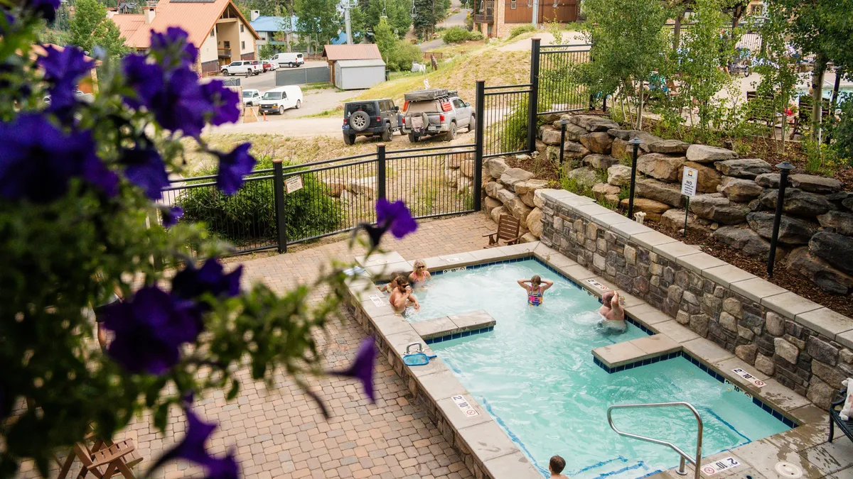 A cheerful exterior shot of a heated outdoor swimming pool surrounded by colorful flowers. Children are seen playing near a rock wall, and the atmosphere is lively and joyful, evoking memories of social gatherings and family relaxation.