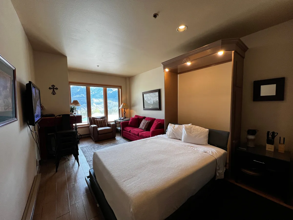 The image showcases a bright and inviting studio condo interior featuring a plush bed with crisp white linens and a cozy red couch. Expansive windows frame stunning views of the snow-capped Needles Mountains, creating a serene backdrop. The space includes a television, stylish side table with decor, and a warm, welcoming atmosphere perfect for relaxation.