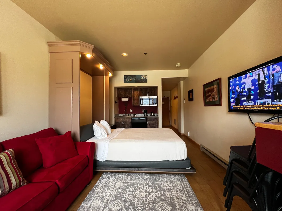 A functional and inviting studio layout, featuring a bright red sofa, a bed for restful sleep, a kitchenette for preparing meals, and a television for entertainment. The space is well-organized for both lounging and practical living, perfect for couples or solo adventurers.