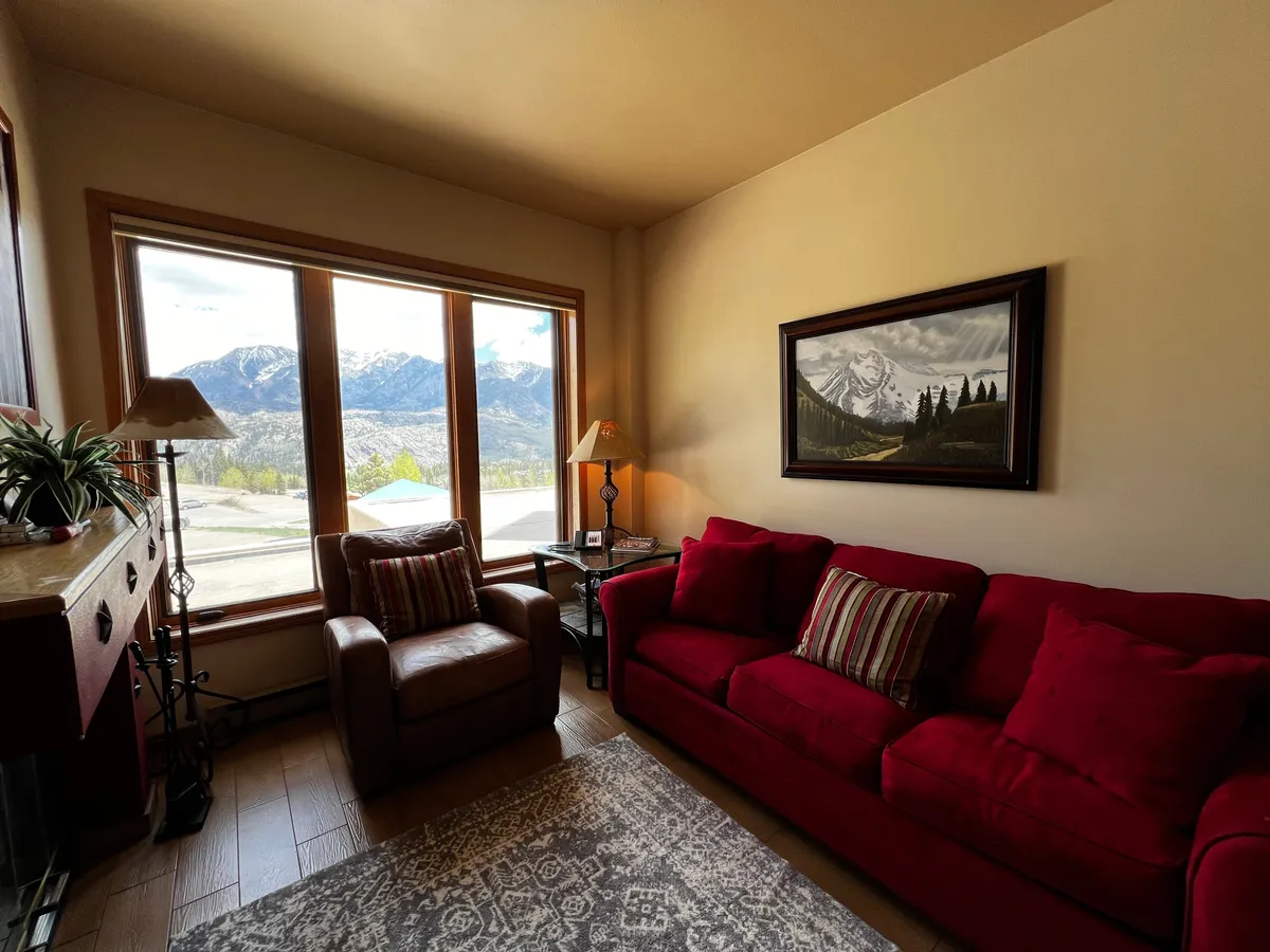 A serene interior corner with a red couch situated by a large window offering mountain views, accented with warm lighting and artwork. The scene invites relaxation, reading, or quiet moments while soaking in the natural surroundings.