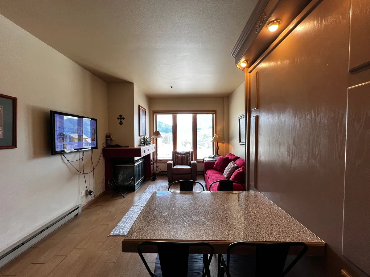 This inviting studio space features a comfortable sofa, a television, expansive windows framing scenic mountain views, a cozy fireplace, and a dining table. The room is bathed in natural light, with the fireplace and windows creating a warm and tranquil atmosphere perfect for relaxation.